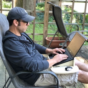 May include: A person sitting in a chair outdoors using a laptop on a lap desk. The laptop is open and the person is typing on the keyboard. The lap desk is made of wood and has a mouse pad on the right side. The person is wearing a black hoodie and a baseball cap.