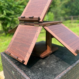 May include: A wooden, foldable stand with a dark brown finish. The stand has multiple levels and is designed to hold books or other items. It is sitting on a wicker table.