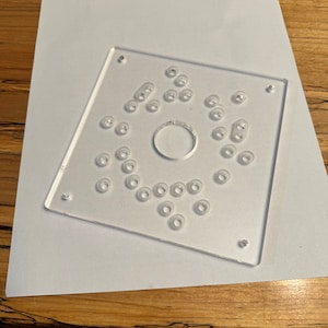 Op de afbeelding: Een transparante plastic sjabloon met een ronde uitsparing in het midden en meerdere kleinere ronde gaten die in een patroon rond de uitsparing zijn gerangschikt.