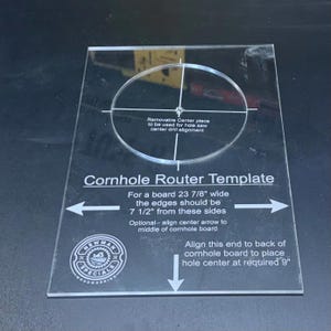May include: A clear acrylic Cornhole Router Template with printed instructions and markings. The template features a circular cutout, a removable center piece, and text specifying alignment for a 23 7/8 inch wide board and 9 inch hole placement.