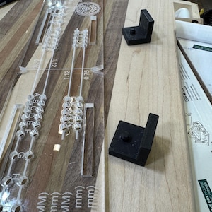 Puede incluir: Una regla de plástico transparente con una serie de agujeros y marcas para medir. La regla está etiquetada con medidas en milímetros. Dos soportes de esquina de plástico negro están sobre una superficie de madera.