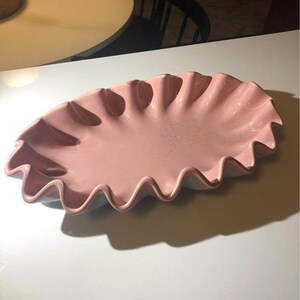 Puede incluir: Una fuente de cerámica rosa con borde ondulado. La fuente, de forma ovalada, está sobre una superficie blanca. El borde ondulado añade un toque decorativo a la fuente.