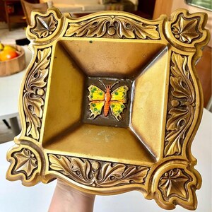 Puede incluir: Una bandeja decorativa de color dorado con un diseño de mariposa en relieve. La mariposa tiene alas amarillas con marcas negras y un cuerpo rojo.