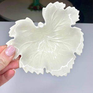 Puede incluir: Un plato de cerámica blanco en forma de hoja con vetas detalladas y un borde festoneado. El plato está sostenido por una mano con una uña pintada de rosa. El plato es probablemente para fines decorativos o de servicio.