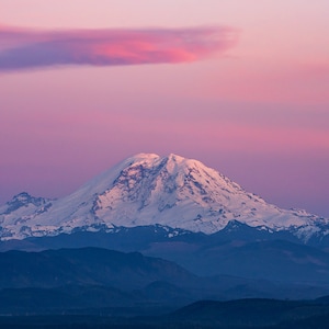以下が含まれることがあります： ピンクと紫の鮮やかな夕焼け空に、雪に覆われた山の頂上がそびえ立っています。山は、低い森林に覆われた丘の連なりで囲まれています。