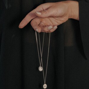 May include: Two silver chain necklaces with round pendants hanging from a hand. The pendants have a textured surface.