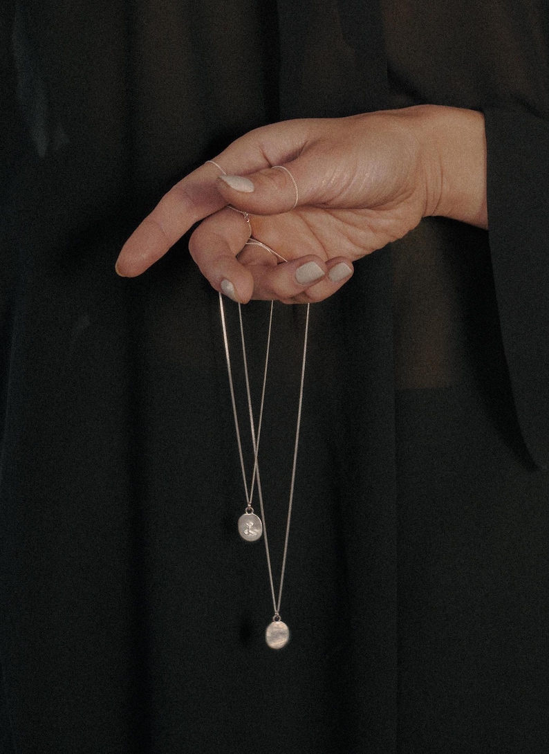 May include: Two silver chain necklaces with round pendants hanging from a hand. The pendants have a textured surface.