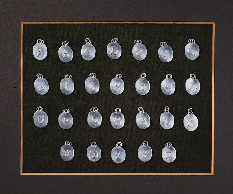 May include: A framed display of 26 silver charms, each featuring a different letter of the alphabet. The charms are arranged in rows and columns on a black velvet background.