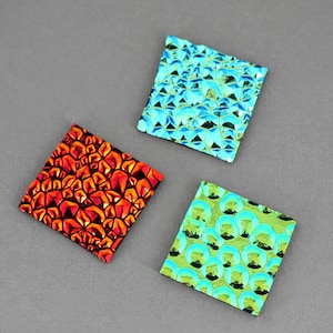 May include: Three square tiles with different iridescent patterns. The first tile is blue with a geometric pattern. The second tile is red with a geometric pattern. The third tile is green with a circular pattern.