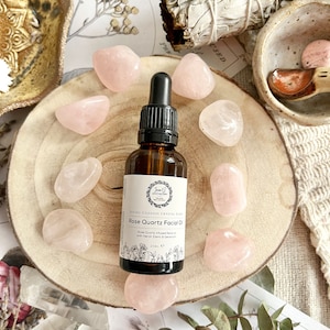 May include: A glass bottle of rose quartz facial oil with a dropper, surrounded by pink rose quartz crystals on a wooden surface. The bottle label reads "Divine Goddess Crystal Elixir Rose Quartz Facial Oil".