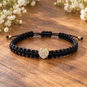May include: A black braided cord bracelet featuring a central gold-toned heart charm encrusted with clear crystals. The adjustable bracelet includes two small gold-toned beads at the ends of the pull-string closure mechanism.