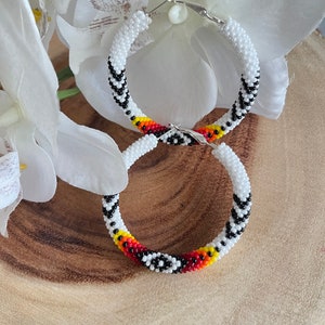 May include: A pair of white beaded hoop earrings with black, red, yellow, and orange accents. The earrings are handmade and feature a geometric pattern.