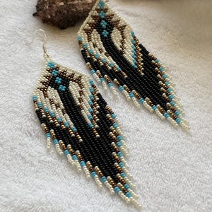 May include: A pair of long, beaded earrings with a geometric pattern in black, white, gold, and turquoise. The earrings are made of seed beads and have a fringe-like design.