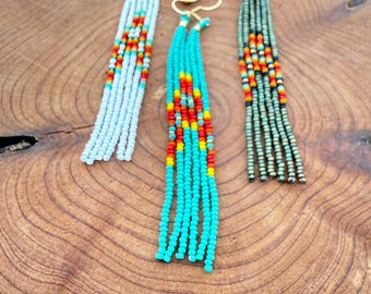 May include: Three pairs of beaded tassel earrings in white, turquoise, and olive green. Each pair has a gold-coloured hook and a pattern of red, orange, and yellow beads. The earrings are displayed on a wooden surface with a starfish.