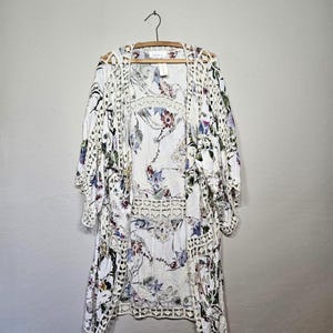 May include: White floral print kimono-style cover-up with openwork crochet trim. The cover-up features a delicate floral pattern in shades of purple, green, and blue. It hangs on a wooden hanger against a neutral background.