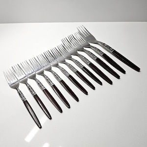 Puede incluir: Un juego de diez tenedores vintage. Cada tenedor presenta una cabeza de acero inoxidable y un mango marrón oscuro con un patrón plateado decorativo. Los tenedores están dispuestos en diagonal sobre una superficie blanca.