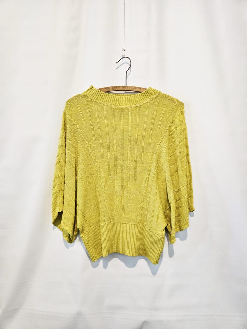 May include: A yellow knitted sweater with a ribbed texture and a round neckline. The sweater has a relaxed fit and short sleeves.