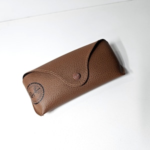 May include: A brown textured leather-like glasses case with a pointed flap and a matching snap button closure. The case features a black circular Ray-Ban logo printed on the lower left corner, indicating the brand of the eyewear accessory.