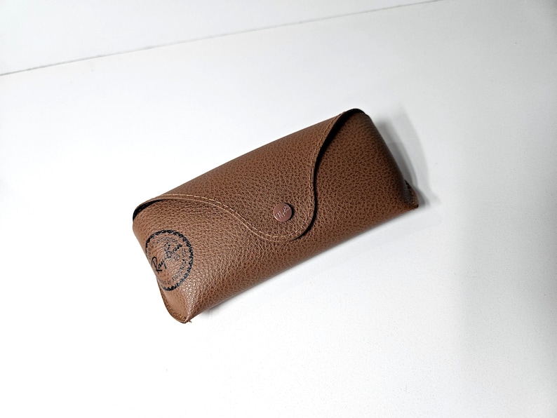 May include: A brown textured leather-like glasses case with a pointed flap and a matching snap button closure. The case features a black circular Ray-Ban logo printed on the lower left corner, indicating the brand of the eyewear accessory.
