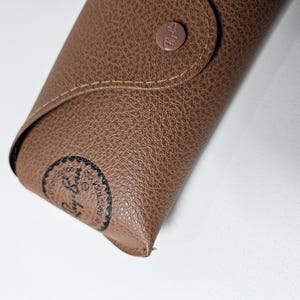May include: A tan, textured faux leather glasses case with a snap button closure. The case features a circular Ray-Ban logo embossed on the front flap and a black circular Ray-Ban stamp on the lower left corner, with visible stitching along the edges.