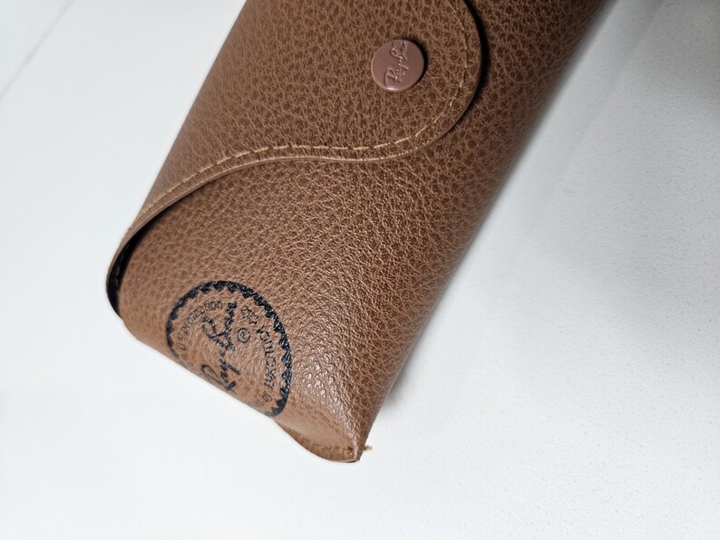 May include: A tan, textured faux leather glasses case with a snap button closure. The case features a circular Ray-Ban logo embossed on the front flap and a black circular Ray-Ban stamp on the lower left corner, with visible stitching along the edges.