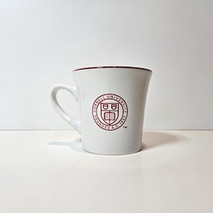 May include: A white ceramic mug with a curved handle and a dark red interior rim. The front features the circular Cornell University seal in dark red, including the text Cornell University Founded A.D. 1865 and a shield emblem with a book.