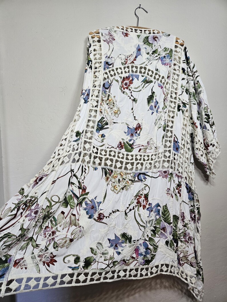 Op de afbeelding: Witte kimono-top met bloemenprint en openwerk gehaakte details. De top heeft een levendig bloemenpatroon in tinten rood, blauw en groen. De mouwen zijn driekwart en het kledingstuk hangt aan een houten hanger.