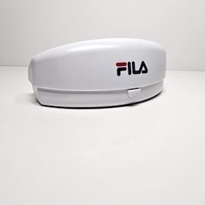 May include: A white, oval-shaped case with the FILA logo in black and red. The case has a hinged lid and a small, rectangular clasp. The product is likely a case for eyeglasses or sunglasses.