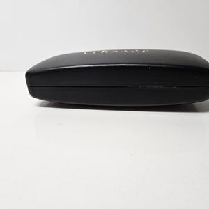 May include: A black, oval-shaped sunglasses case with the brand name "VERSACE" in gold lettering on the top. The case is closed and appears to be made of a smooth material.