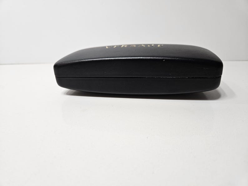May include: A black, oval-shaped sunglasses case with the brand name "VERSACE" in gold lettering on the top. The case is closed and appears to be made of a smooth material.