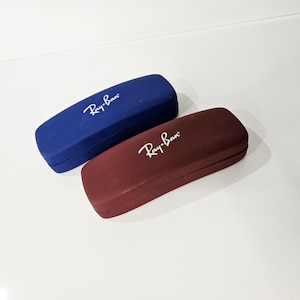 Puede incluir: Dos estuches rectangulares para gafas. Uno es azul oscuro, el otro de color burdeos. Ambos estuches tienen el logotipo de Ray-Ban en escritura blanca. Los estuches están sobre una superficie blanca.