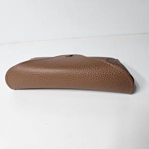 May include: A brown, textured, semi-rigid glasses case with a curved, envelope-style flap closure. The case features a pebbled surface finish and a single central snap button fastener, presenting a sleek, compact, and rounded rectangular shape.