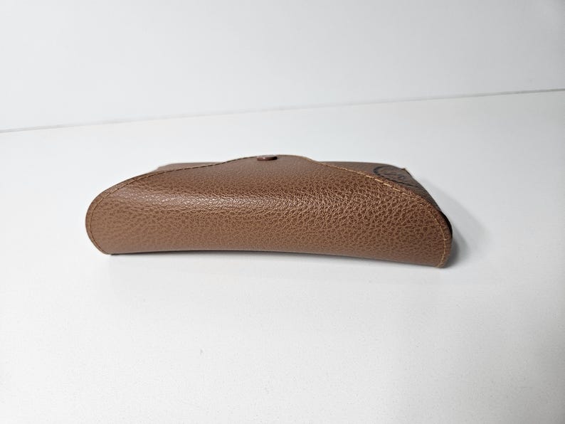 May include: A brown, textured, semi-rigid glasses case with a curved, envelope-style flap closure. The case features a pebbled surface finish and a single central snap button fastener, presenting a sleek, compact, and rounded rectangular shape.