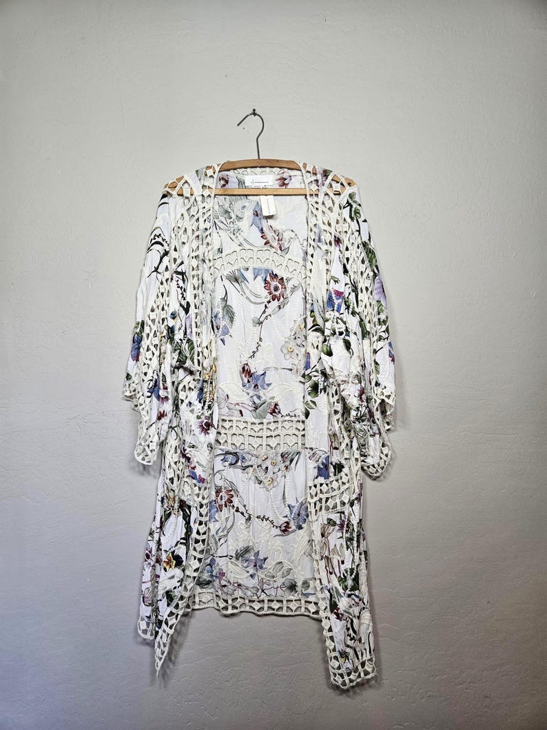 Op de afbeelding: Witte kimono-achtige cover-up met bloemenprint en openwerk gehaakte rand. De cover-up heeft een delicaat bloemenpatroon in tinten paars, groen en blauw. Het hangt aan een houten hanger tegen een neutrale achtergrond.