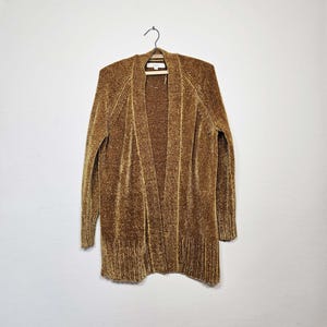 May include: A long, open-front cardigan in a warm, golden-brown hue hangs on a wooden hanger. The sweater has a ribbed texture and a soft, plush appearance. The cardigan's length extends to the mid-thigh, with long sleeves and a relaxed fit.