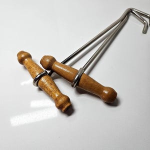 May include: Two metal hooks with T-shaped wooden handles. Each handle features a turned, cylindrical design with a smooth, light brown finish. The metal shafts are silver-colored and extend from the center of each handle, ending in a curved hook.