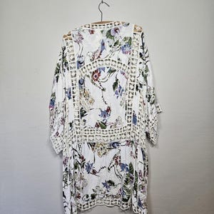 Op de afbeelding: Witte kimono-top met bloemenprint en openwerkdetails. De top heeft een V-hals en driekwartmouwen. Het bloemenpatroon bevat blauwe, paarse en rode bloemen op een witte achtergrond. Het kledingstuk hangt aan een houten hanger.