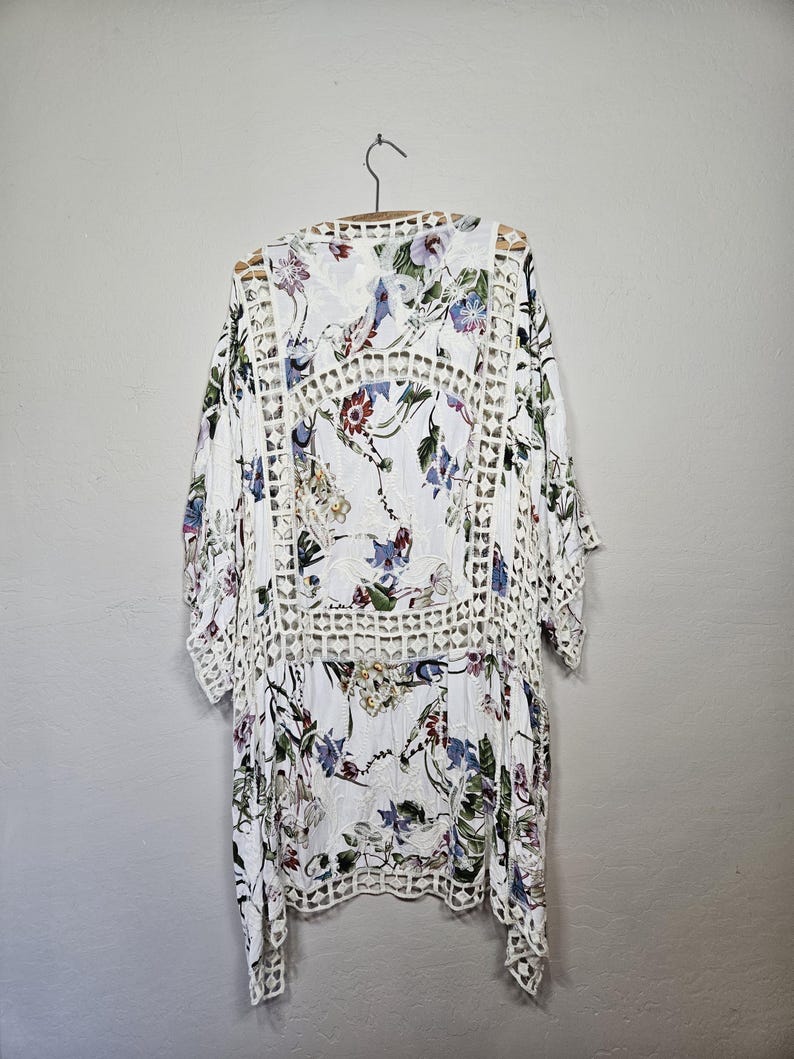 Op de afbeelding: Witte kimono-top met bloemenprint en openwerkdetails. De top heeft een V-hals en driekwartmouwen. Het bloemenpatroon bevat blauwe, paarse en rode bloemen op een witte achtergrond. Het kledingstuk hangt aan een houten hanger.