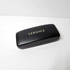 May include: A black rectangular glasses case with the gold-colored word "VERSACE" printed on the top. The case is closed and appears to be made of leather or a similar material. The case is on a white surface.