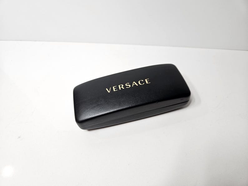 May include: A black rectangular glasses case with the gold-colored word "VERSACE" printed on the top. The case is closed and appears to be made of leather or a similar material. The case is on a white surface.