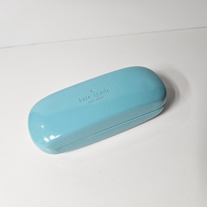 May include: A light blue, hard-shell, rectangular glasses case with rounded edges. The top surface features the embossed text 'kate spade NEW YORK' along with a small spade logo symbol centered on the lid of the closed protective container.