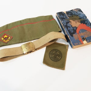 May include: An assortment of vintage Boy Scout items, including a green cap with a red trim, a tan belt with a gold buckle, a patch, and a well-worn "Handbook for Boys" book.