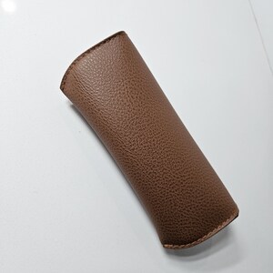 May include: A brown, textured, semi-rigid slip-in case with a curved top opening and visible stitching along the edges. The exterior features a pebbled grain finish, providing a tactile surface for this protective sleeve designed for eyewear.