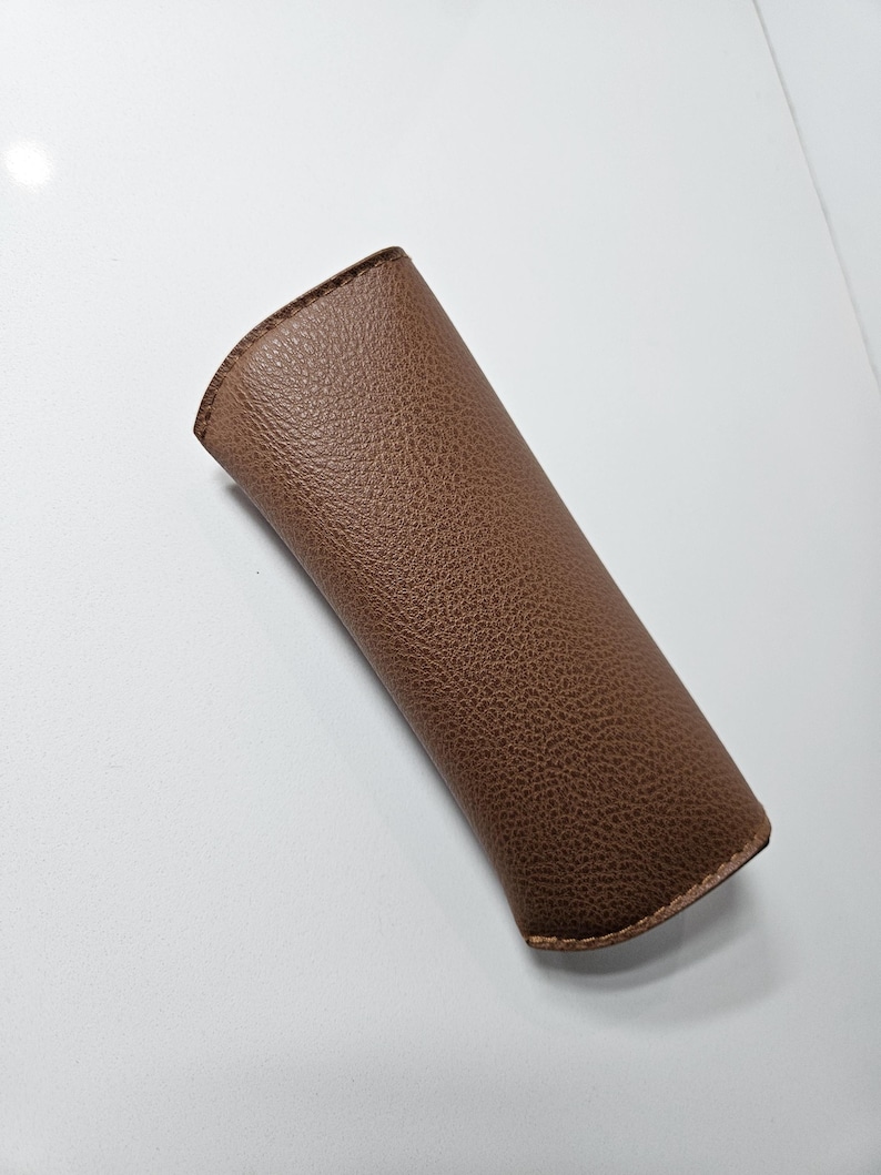 May include: A brown, textured, semi-rigid slip-in case with a curved top opening and visible stitching along the edges. The exterior features a pebbled grain finish, providing a tactile surface for this protective sleeve designed for eyewear.