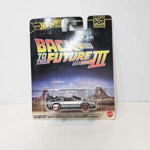 Puede incluir: Un coche de juguete Hot Wheels Premium Pop Culture, modelo Back to the Future Part III Time Machine 50's Version. El vehículo plateado está dentro de su embalaje original de cartón negro con el título y el logotipo de la película.