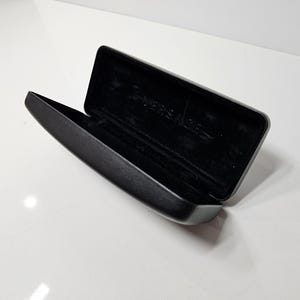 May include: An open, black rectangular glasses case with a soft, dark interior. The case is made of a smooth, matte material and features the brand name "VERSACE" embossed inside. The case is set against a white background.