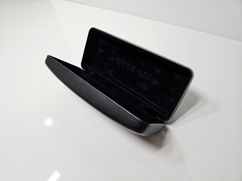 May include: An open, black rectangular glasses case with a soft, dark interior. The case is made of a smooth, matte material and features the brand name "VERSACE" embossed inside. The case is set against a white background.