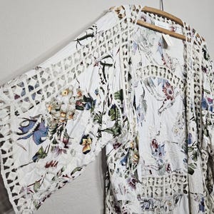 Op de afbeelding: Witte bloemenkimono met openwerk kanten afwerking. De kimono heeft een bloemenpatroon in tinten blauw, paars, groen en geel. De mouwen en randen hebben een geometrisch kantontwerp. Het kledingstuk hangt aan een houten hanger.