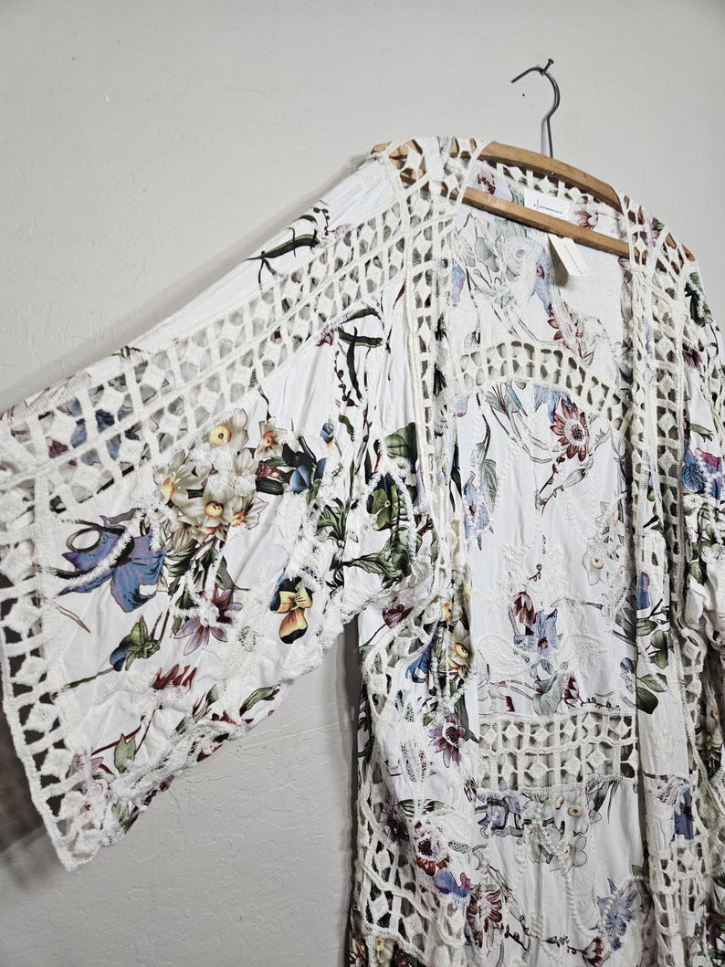 Op de afbeelding: Witte bloemenkimono met openwerk kanten afwerking. De kimono heeft een bloemenpatroon in tinten blauw, paars, groen en geel. De mouwen en randen hebben een geometrisch kantontwerp. Het kledingstuk hangt aan een houten hanger.