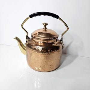 May include: A copper tea kettle with a black handle and brass accents. The kettle has a hammered texture and a brass spout. The lid is also copper and has a small knob. The handle is arched and attached to the kettle with brass brackets.